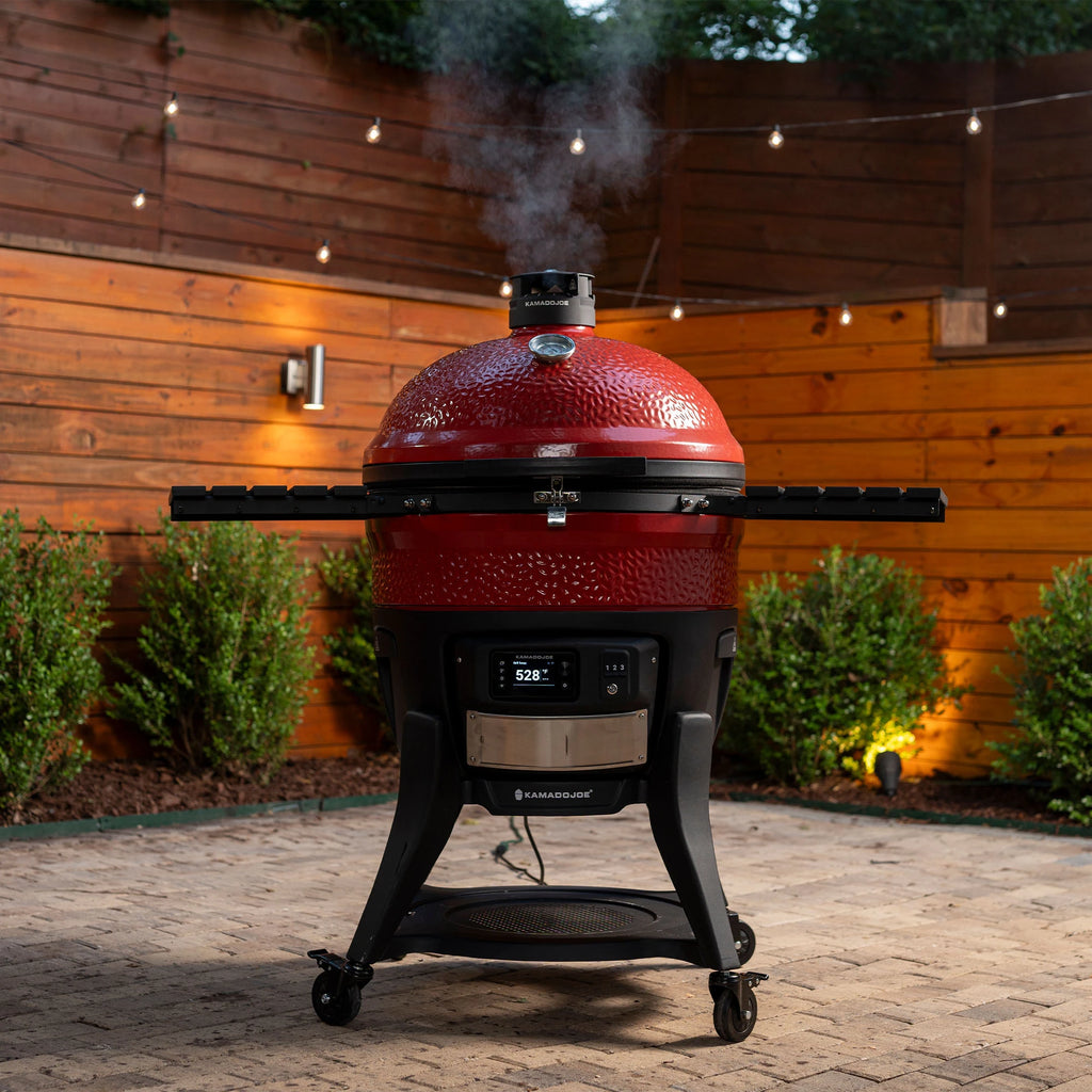 A Big Joe Konnected Joe Digital Charcoal Grill and Smoker outside on a brick patio in front of bushes and 2 stained wood fences. Smoke escapes from the Kontrol Tower top vent.