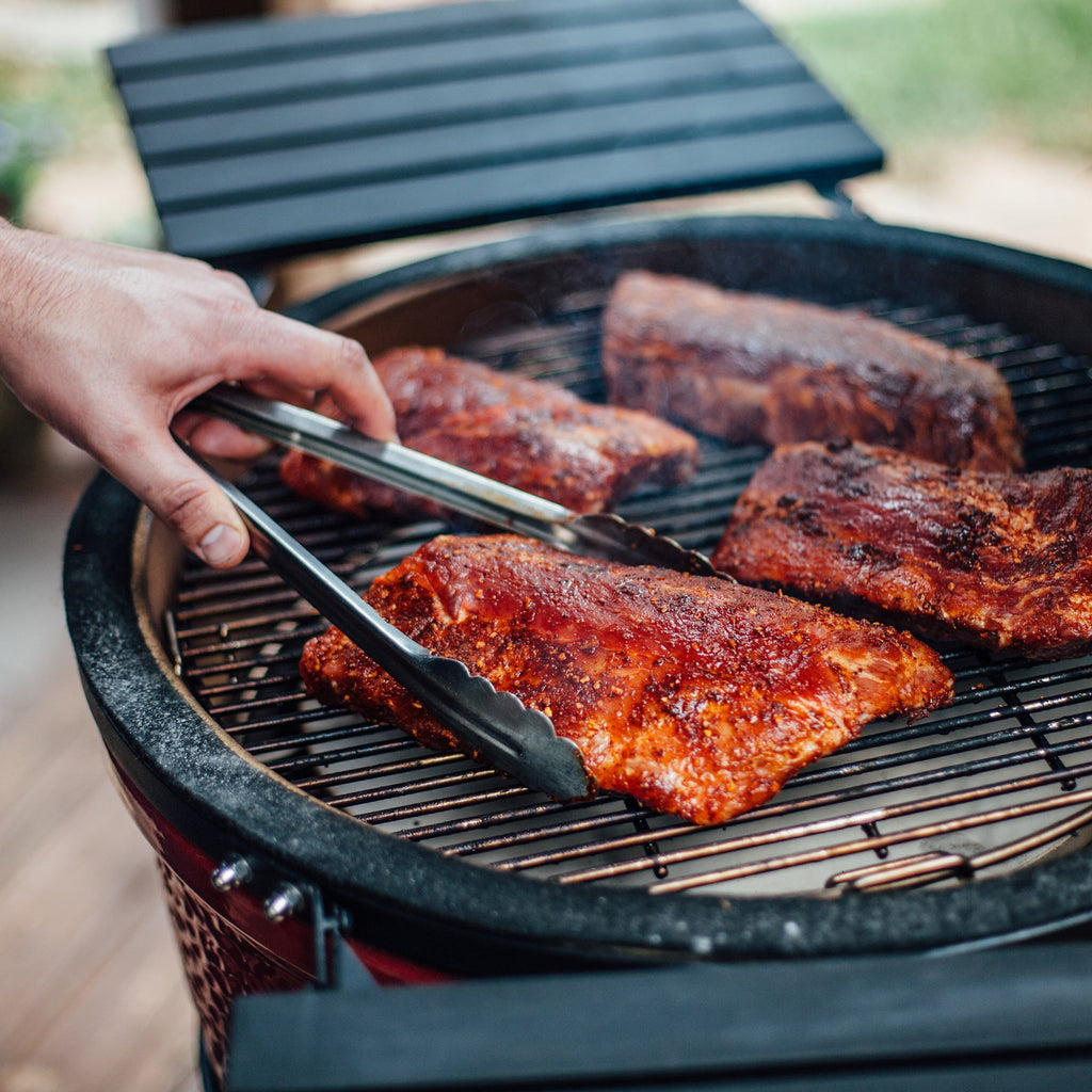 A hand holding tongs come in frame from the left, using the tongs to grip a seasoned rack of ribs cooking on the grate over a heat deflector. The grill's side shelves are also visible.
