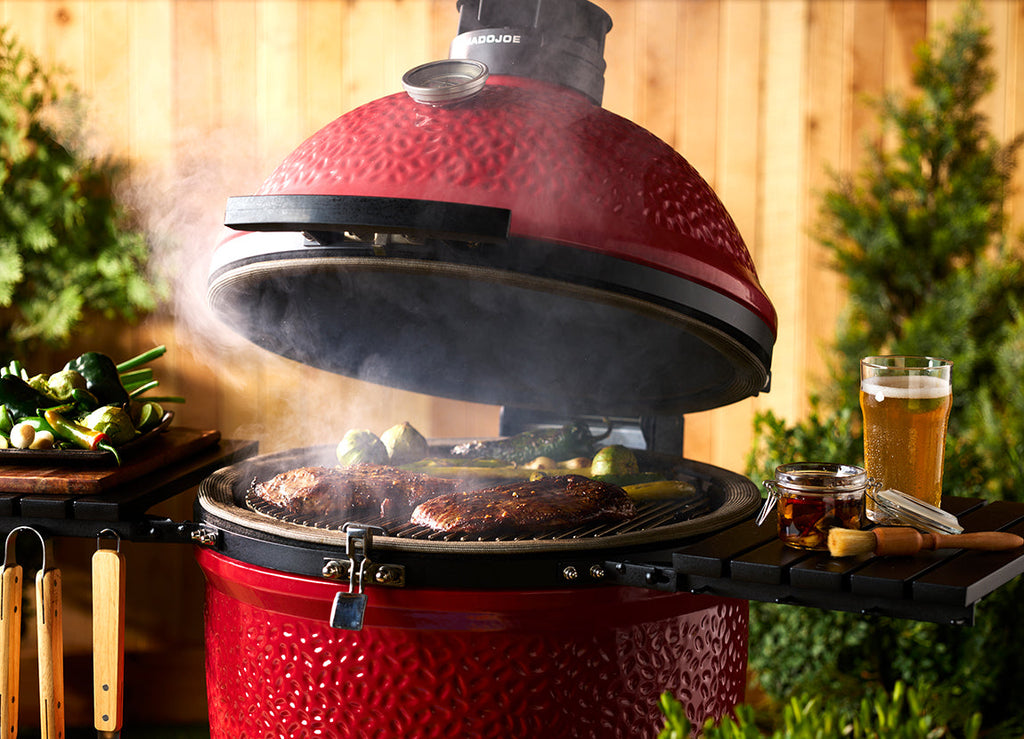 A partially opened grill with smoke rising around steaks and vegetables cooking on the grill. Both side shelves are in use and wooden-handled utensils hang from the hooks on the left shelf