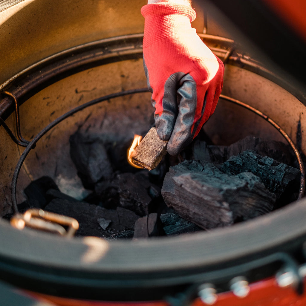 Someone wearing an orange and black grill glove reaches into an open Kamado Joe grill to place a lit fire starter on top of a pile of big blog XL lump charcoal.