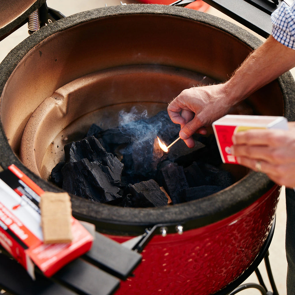 A man reaches into an open Kamado Joe grill as he holds a lit match near the pile of XL Big Block  charcoal in the bottom of the grill. 