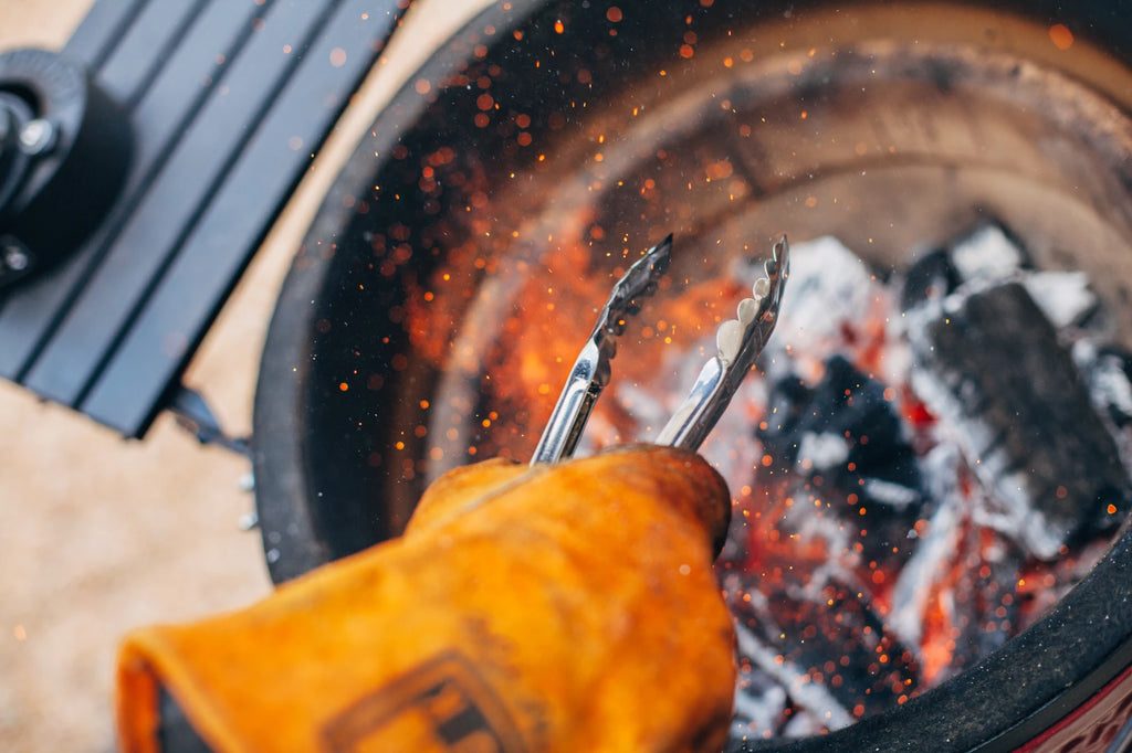 Someone wearing a yellow leather gill glove uses metal tongs to rearrange a pile of lit Big Block XL charcoal as sparks fly up around his hand.