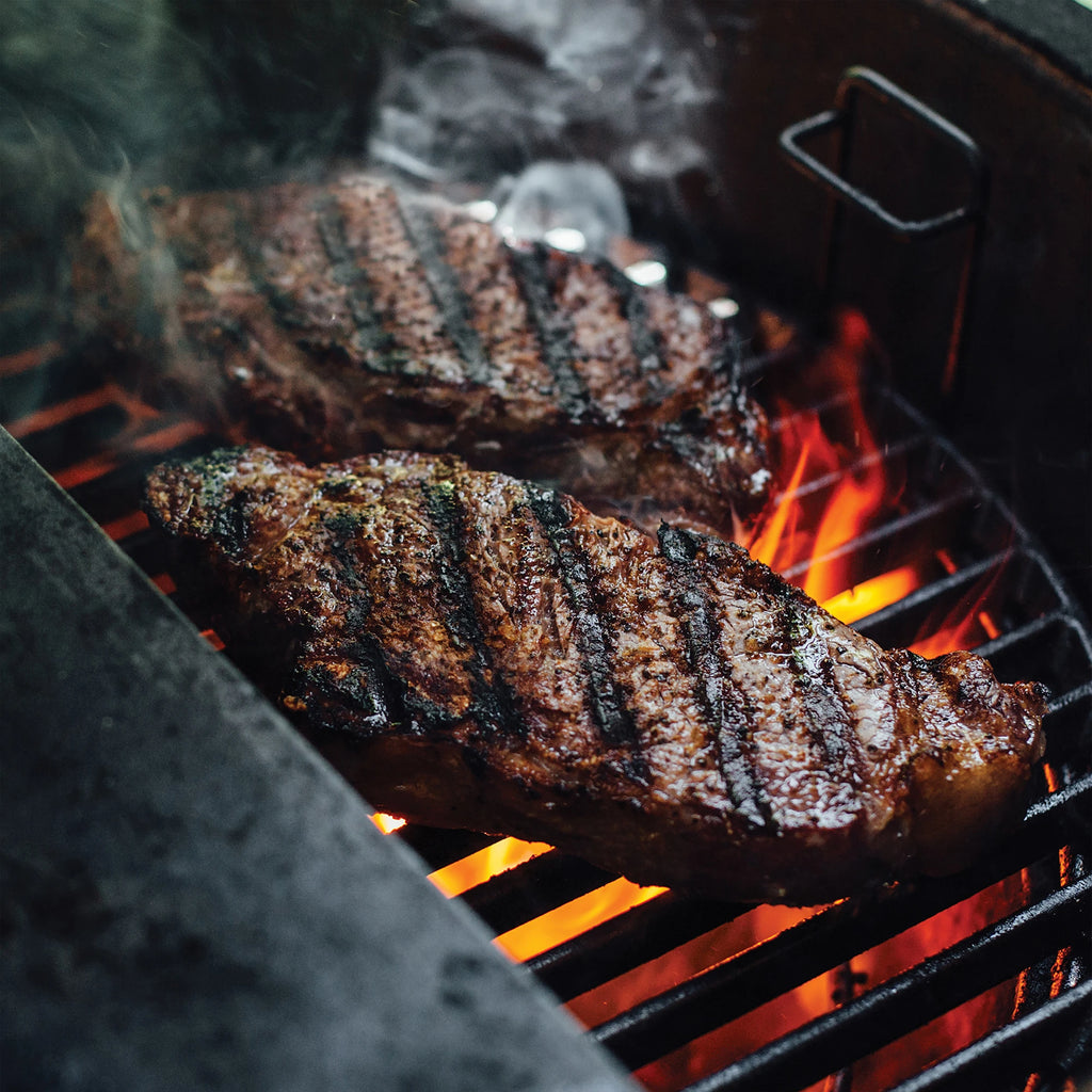 Closeup of seared steaks with dark grill marks on them cooking over the flames coming up through a half-moon cast iron grill grate