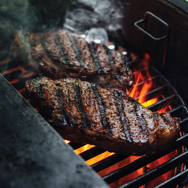 Closeup of seared steaks with dark grill marks on them cooking over the flames coming up through a half-moon cast iron grill grate