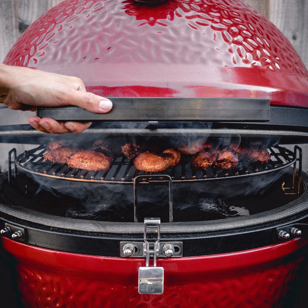 Someone opens a Kamado Joe grill with a SloRoller inside. Chicken wings are cooking on the grates with smoke billowing around them.