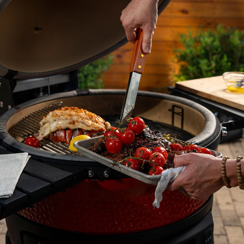 Someone uses tongs to remove grilled tomatoes on the vine, steak, and lobster from the Divide & Conquer cooking racks in a Big Joe Konnected Joe grill