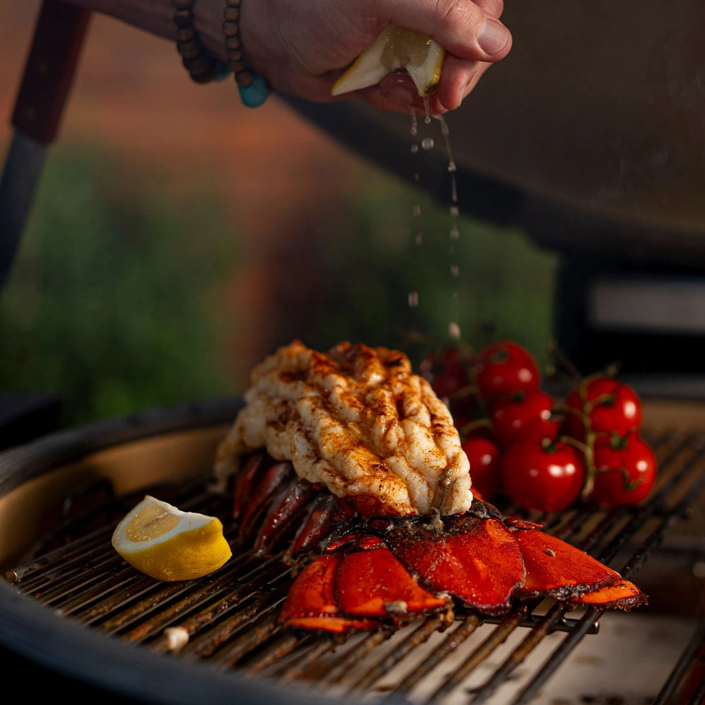 Someone squeezes a fresh lemon over a split and seasoned lobster tail cooking on the grill next to tomatoes on the vine.