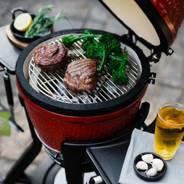 An open Joe Jr grill with 2 round steaks and some broccoli rabe cooking on it. The side shelves hold a drink, a small dish of garlic cloves, and a small dish of fresh herbs with half a lemon.