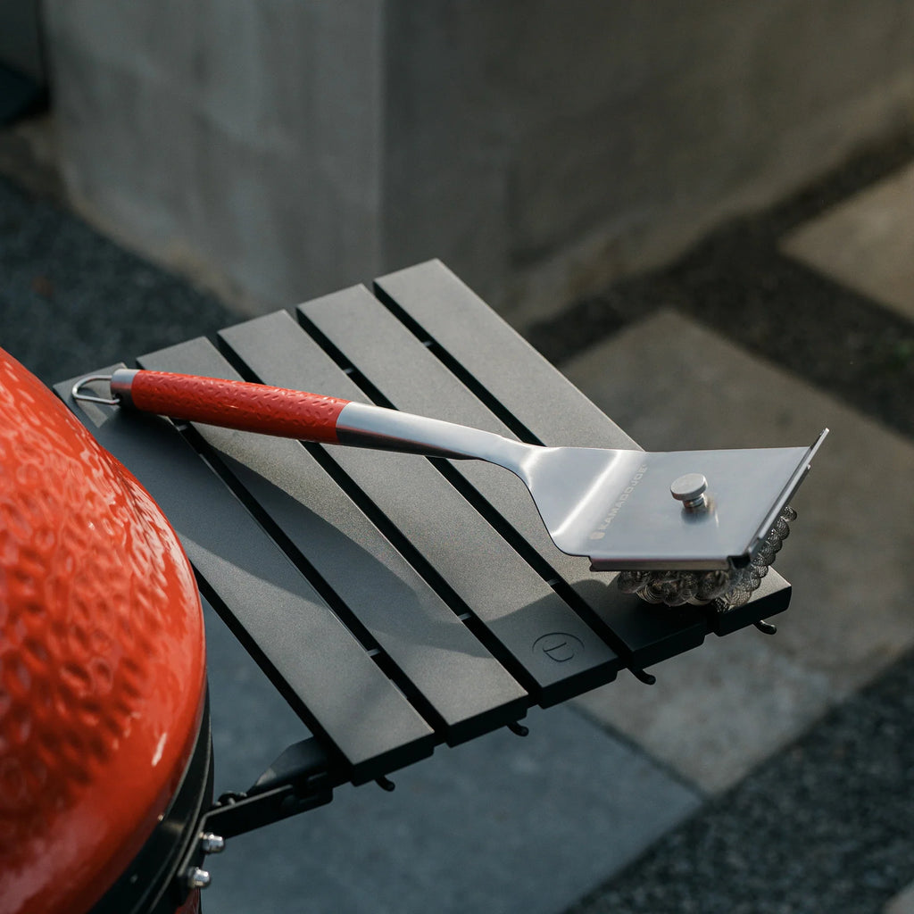 The Kamado Joe stainless steel grill brush resting on the side shelf of a Kamado Joe grill. The bristle side is down to show a small knob on the back of the brush. 