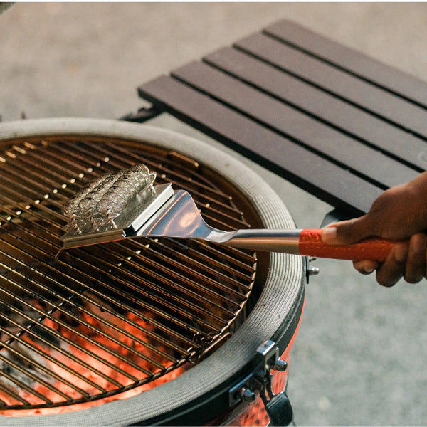 A person uses the built-in grill scraper at the end of a Kamado Joe grill brush to clean a cooking grate in a Kamado Joe grill