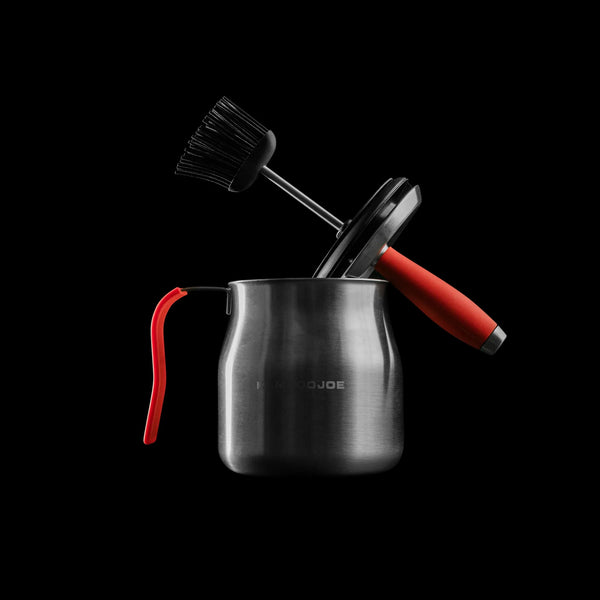 Steel basting pot with red handle cover against a black background. The combined brush and sealing lid with red handle rests on the edge of the pot away from the handle.