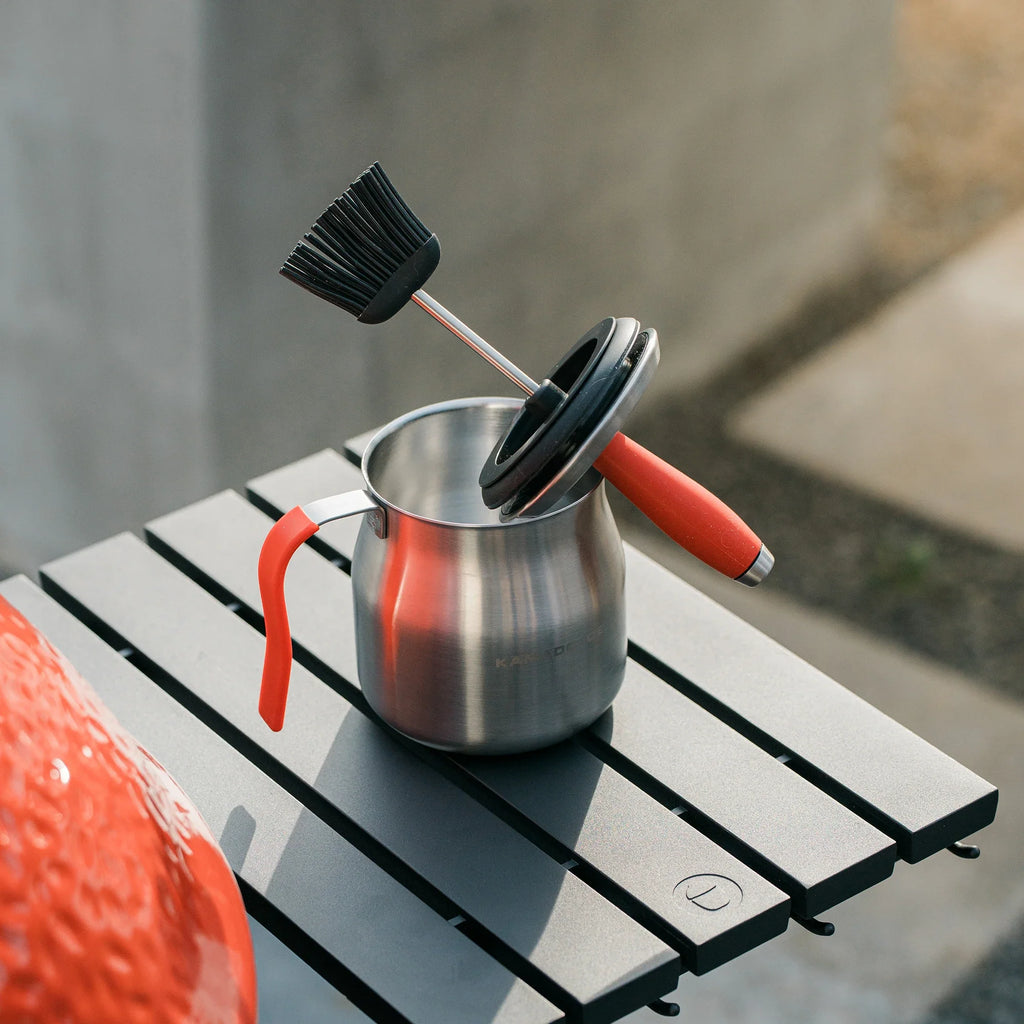 The Kamado Joe basting pot sitting on the side shelf of a Kamado Joe grill. The brush, which has the pot lid mounted on the handle, rests brush up with the pot lid sideways in the pot.