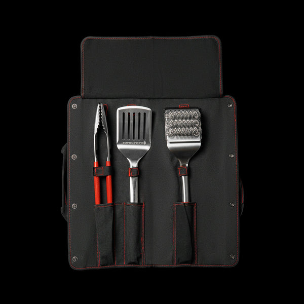 The inside of the canvas case showing the pockets for three tools. From left to right: tongs, spatula, and bristle brush. The case has red accent stitching around the edges, the pockets, and the loop that keep the tools in place. There is a flap attached to the top of the case that folds down over the heads of the tools to protect them when the case is folded for carrying.