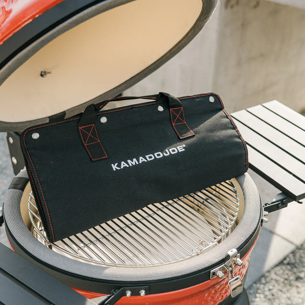 The folded carry case of a Kamado Joe tool kit resting on the grate of an open Kamado Joe grill. The case is black with orange stitching around the edges and where the handles are attached. The words Kamado Joe are embroidered in white on the side of the case.