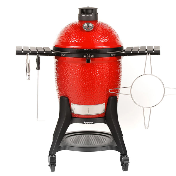 A red Classic Joe III grill with black side shelves, Kontrol Tower top vent, and heavy-duty cart with wheels. A temperature gauge is built into the grill dome. A sliding steel access door is built into the bottom of the grill base. Various accessories hang from the side shelves